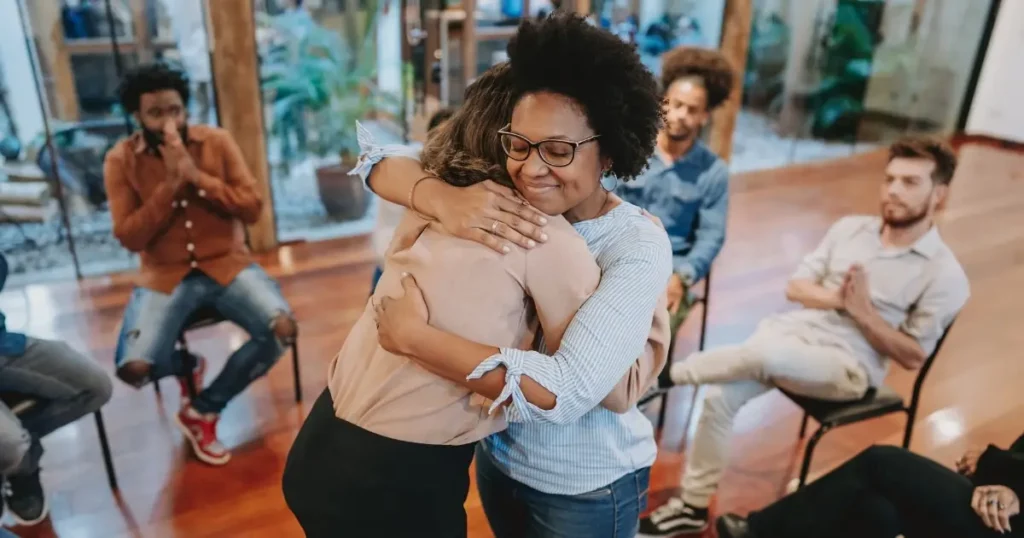 A woman warmly hugging another in a support group session, with people sitting in a safe circle, showing emotional support and healing for anxiety